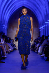 Model walking a runway in a cobalt blue, crocheted high-neck midi dress with feathered blue shoes under blue-lit arches.