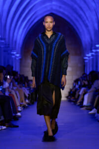 Model walking a runway in a cobalt-blue knitted outer layer over a black blouse and skirt, blue-lit arches in the background.