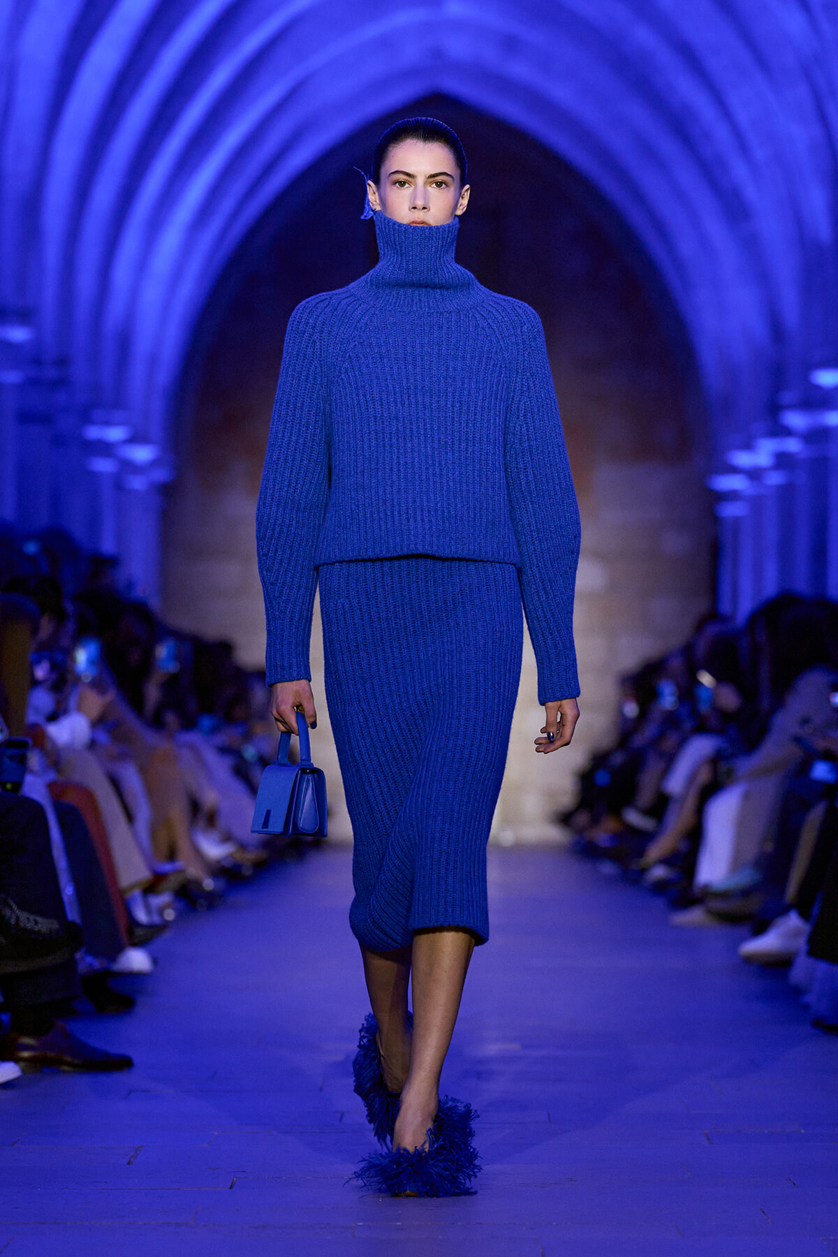 Model walks a runway in a cobalt blue ribbed knit turtleneck and matching midi skirt, carrying a small blue handbag under blue lighting.