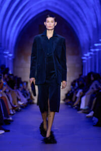 Model walks a fashion show in a navy velvet suit with blazer and matching pants under blue arch lighting on a runway.