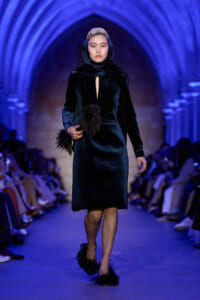 Model walking a fashion show in a black velvet dress with a hooded scarf, blue-lit arches in the background.]”,
