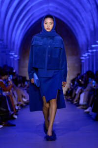 Model walks a runway in a cobalt blue ensemble: oversized jacket with large pockets, matching skirt, and a blue scarf, under blue-lit arches onlookers blurred in background.