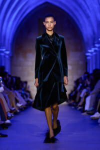 Female model in a dark velvet tailored suit walking a blue-lit runway, wearing a double-breasted blazer and asymmetrical skirt with feathered heels.