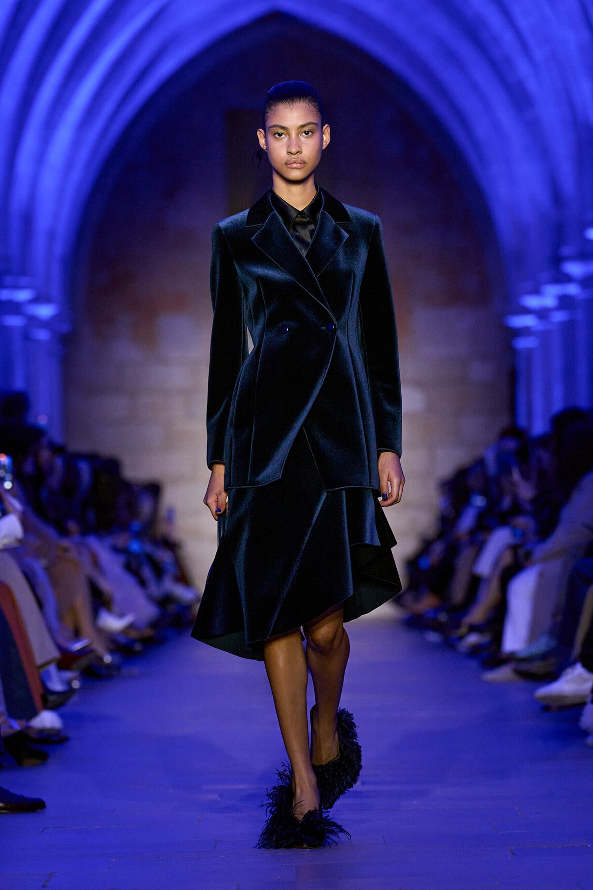Female model in a dark velvet tailored suit walking a blue-lit runway, wearing a double-breasted blazer and asymmetrical skirt with feathered heels.