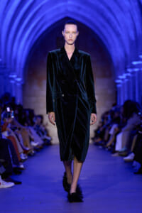 Model walking a runway in a black velvet double-breasted coat under blue arches.