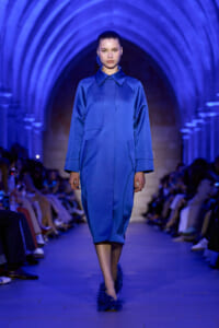 Model walking a fashion runway in a cobalt blue midi coat under blue-lit arches, audience on either side.