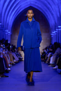 Model walks a blue-coated outfit down a fashion show runway, holding a matching blue clutch under blue lighting.