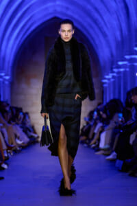Model walking the runway in a dark plaid suit with a black fur vest, carrying a structured black handbag under blue arches.