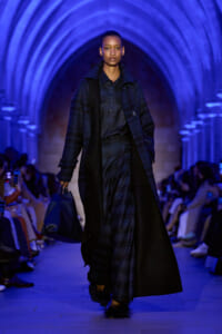 Model walks the runway in a dark blue plaid coat and matching dress, carrying a large navy bag under blue arches.