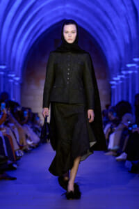 Female model in a dark tailored coat with a buttoned front and cape hood walking a blue-lit runway, carrying a small black handbag.