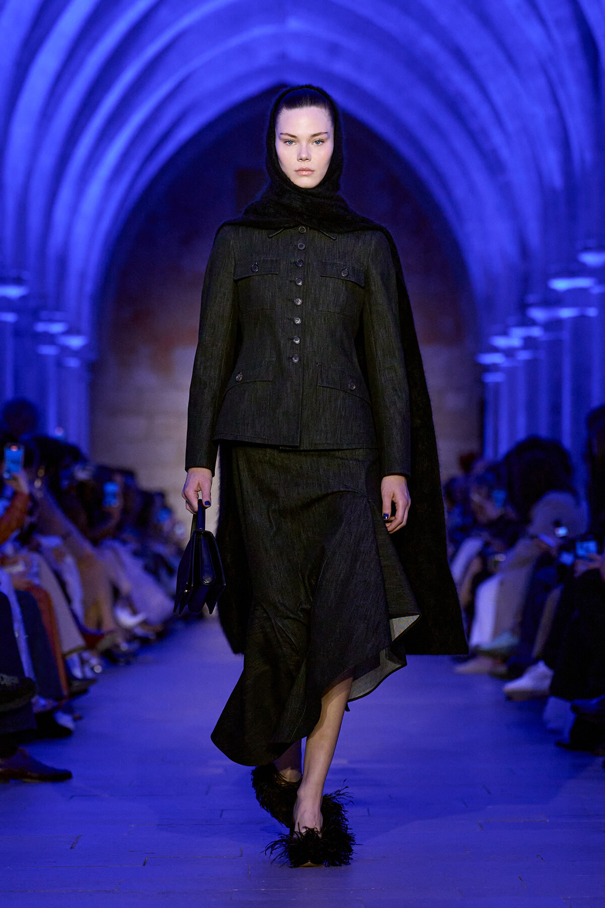Female model in a dark tailored coat with a buttoned front and cape hood walking a blue-lit runway, carrying a small black handbag.