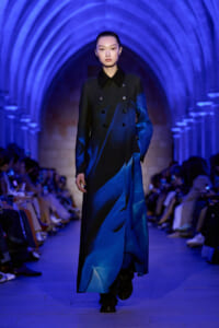 Female model walks the runway in a long, dark-to-blue gradient double-breasted coat under blue arched lighting in a vaulted venue.