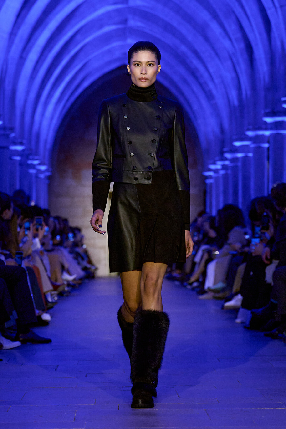 Model walks a blue-lit runway in a black leather double-breasted jacket, matching skirt, and fur boots.