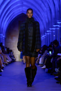 Model walks down a blue-lit runway in a dark plaid outfit and furry boots, holding a clutch.