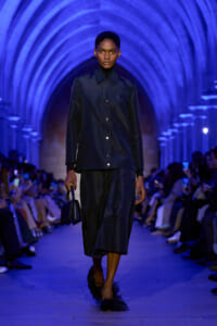 Model walks a blue-lit runway in a dark blue jacket and wide-leg pants, carrying a small matching bag amid arched tunnel set.