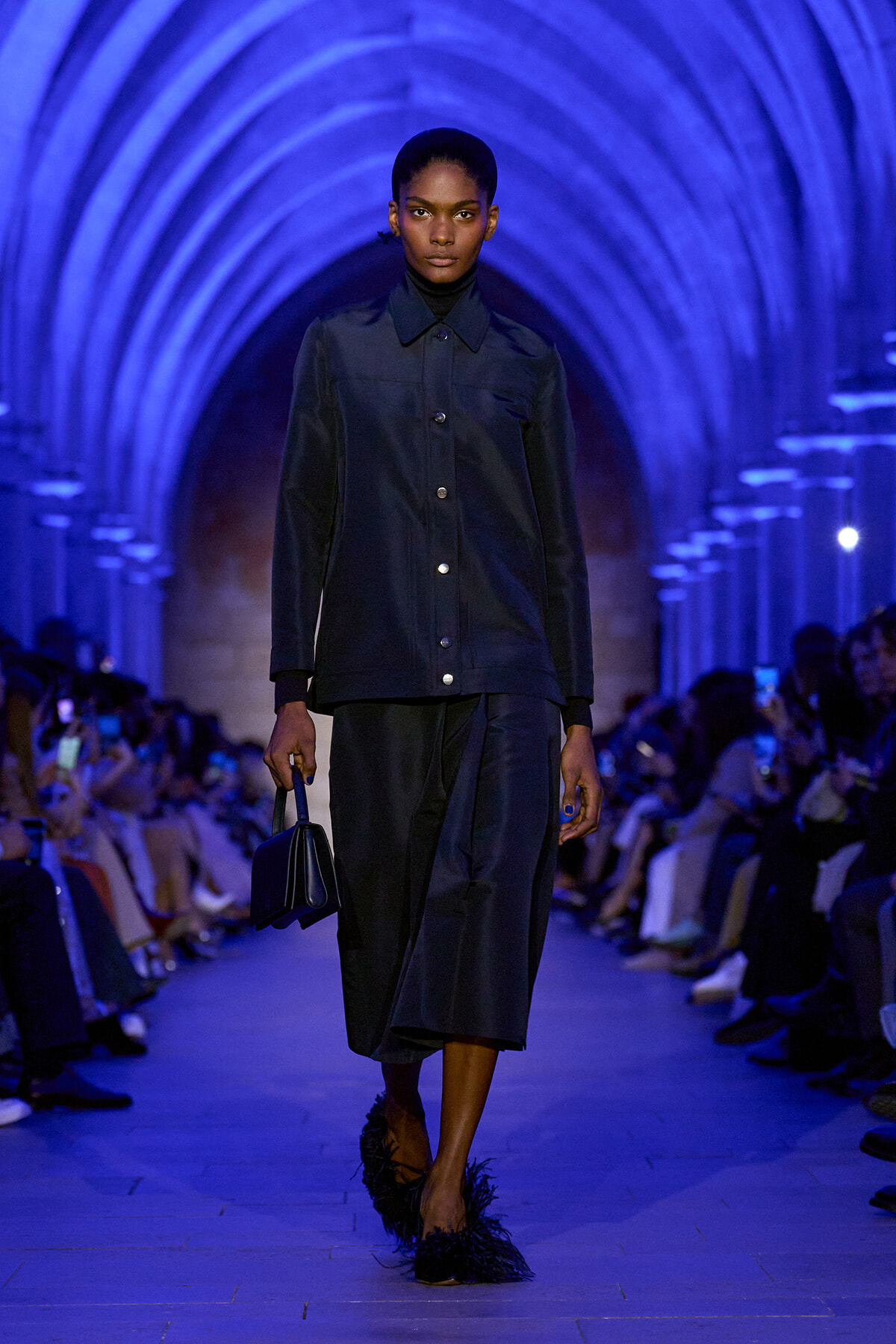 Model walks a blue-lit runway in a dark blue jacket and wide-leg pants, carrying a small matching bag amid arched tunnel set.