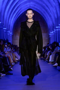 Model walks the runway in a black belted coat with a sheer neckline and layered skirt, under blue lighting in an arched venue.