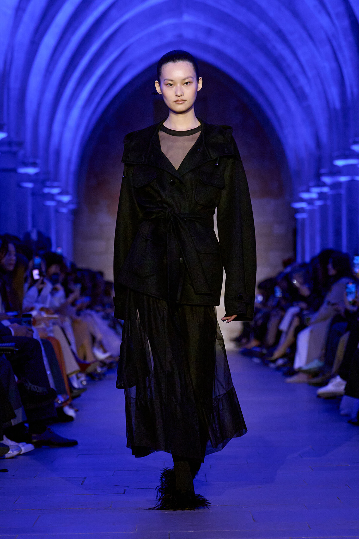 Model walks the runway in a black belted coat with a sheer neckline and layered skirt, under blue lighting in an arched venue.