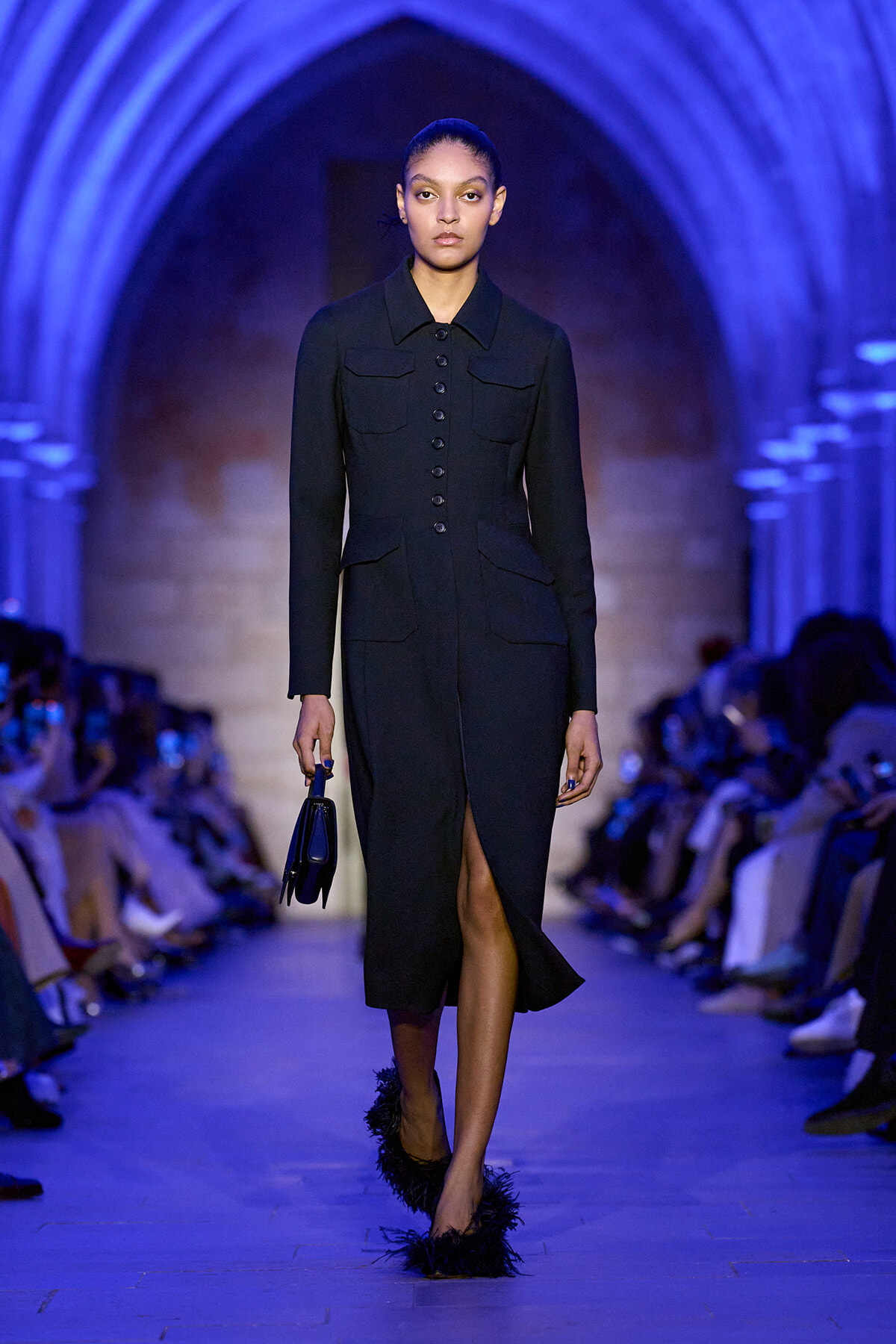 Model walking a dark tailored midi dress with front buttons and pockets on a blue-lit runway archway, carrying a small handbag.