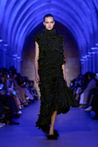 Model walks the runway in a black, ruffled high-neck gown under blue-lit arches.