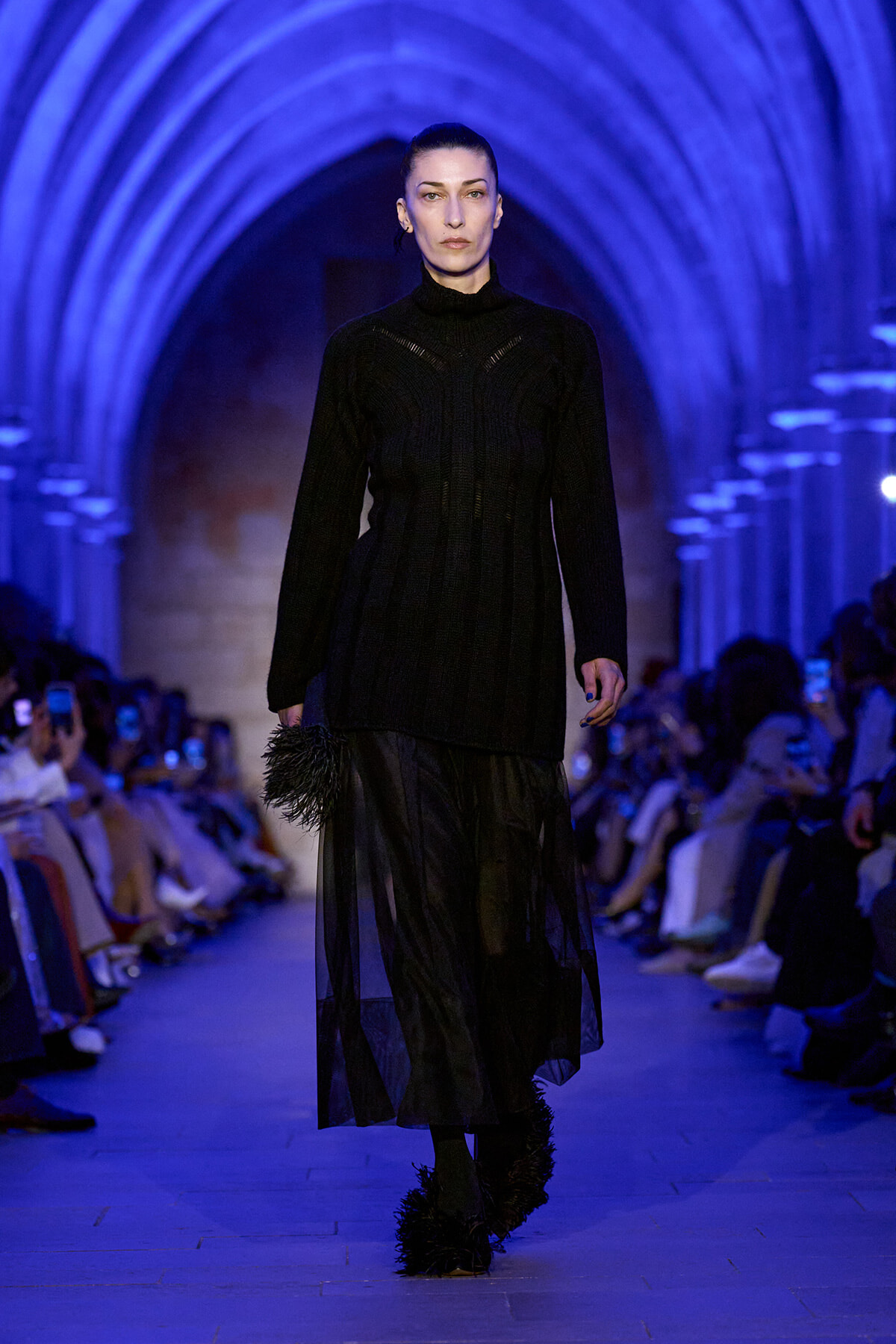 Female model walks a runway in a long black knit dress with sheer panels, in a blue-lit arched venue.
