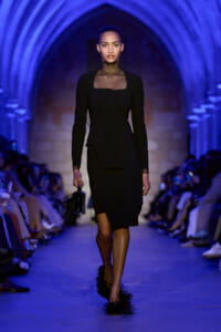 Model walks a blue-lit runway in a fitted black dress with a sheer neckline and long sleeves, carrying a small feathered clutch and wearing feathered heels.