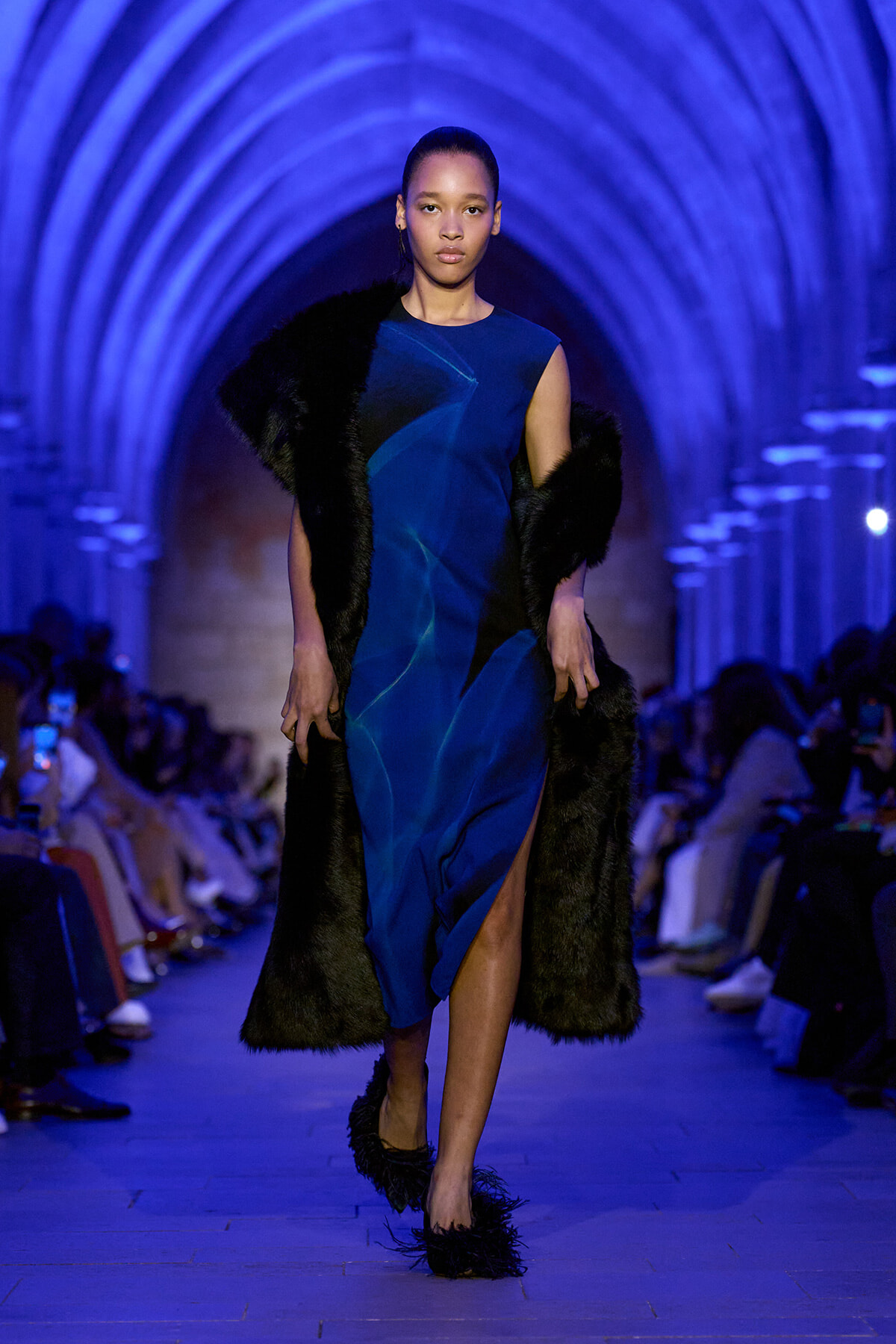 Model walking a cobalt blue dress with a long black fur coat on a blue-lit runway.