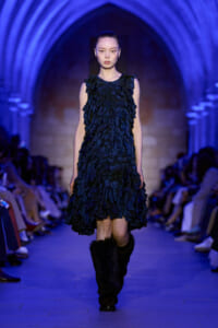 Runway model wearing a navy blue, petal-fringed dress with black fur boots under blue architectural arches.