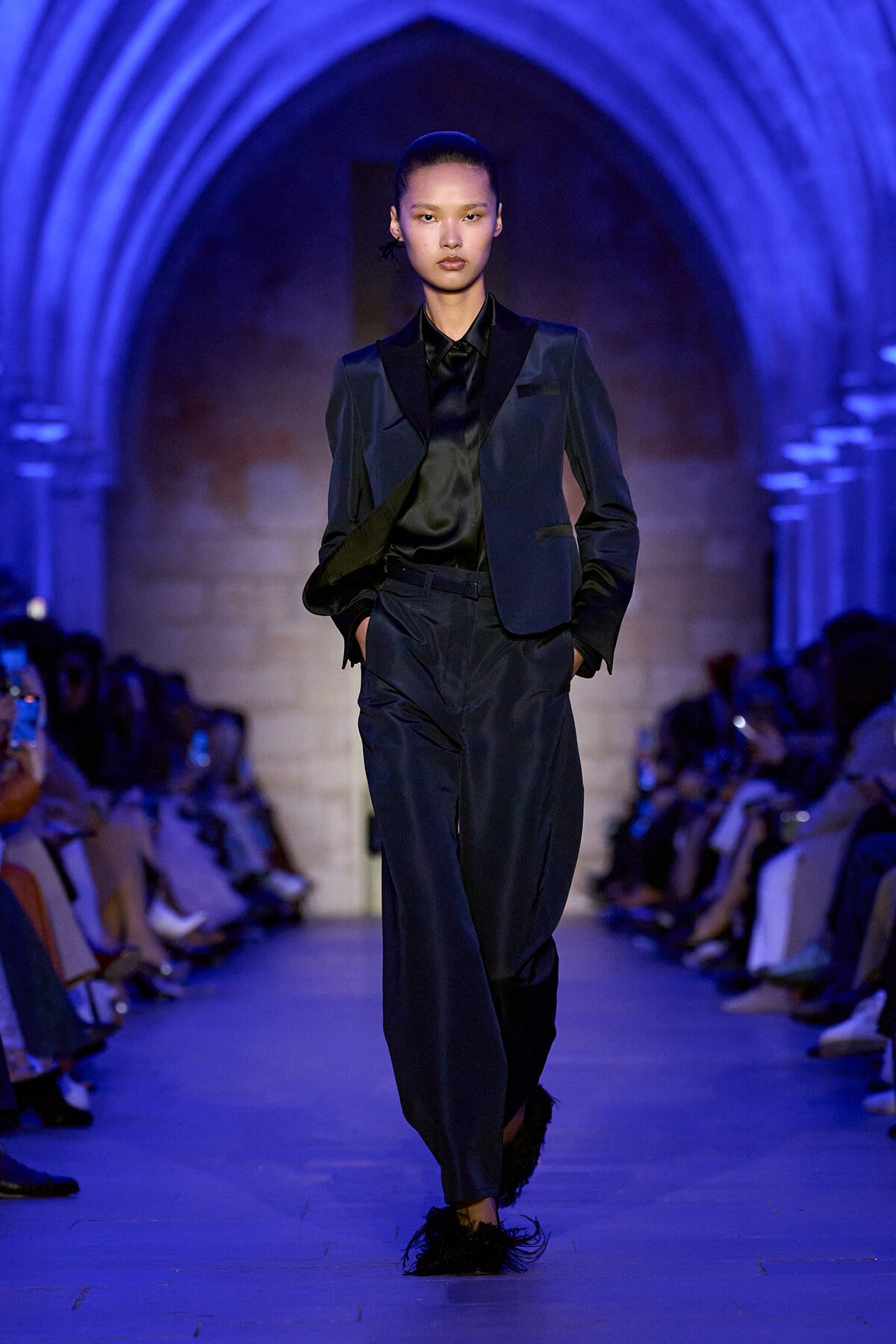 Model walking the runway in a dark navy blazer and matching trousers under blue lighting in a stone-arched venue, hands in pockets.