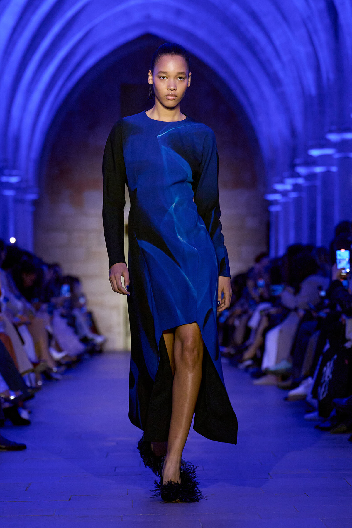 Model walking a runway in a blue and black long-sleeve dress inside a blue-lit arched corridor.