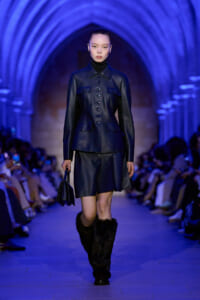 Model walks the runway in a navy leather tailored jacket with matching shorts, black turtleneck, and fur-lined boots under blue lighting. She carries a small dark handbag as spectators watch.