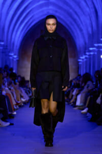Model walks a blue-lit runway under arched ceilings in a black tailored coat, scarf, and fur-trim boots, holding a small black handbag.