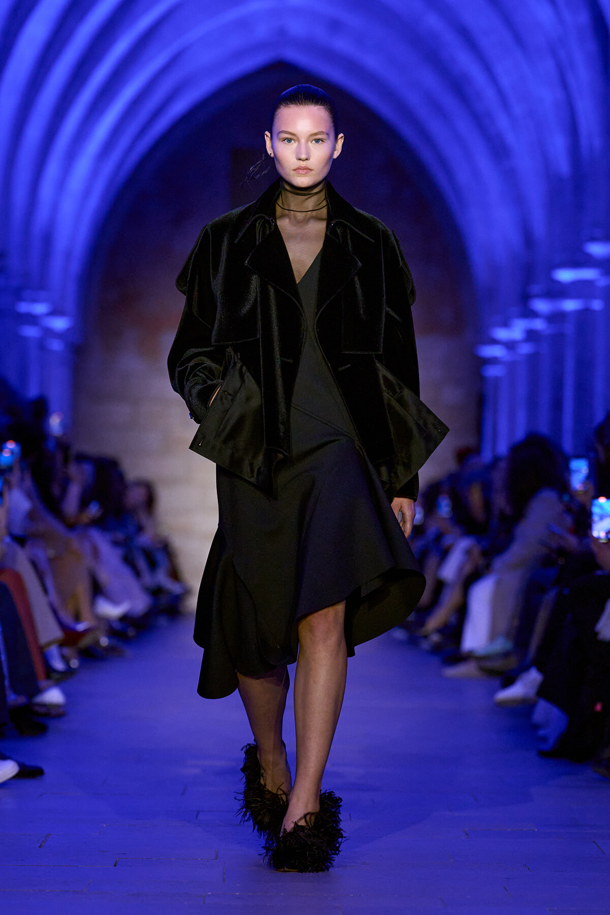 Model walking a runway in a black velvet coat over a black dress, with feathered black shoes, under blue-lit arched ceiling.