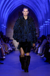 Model walks a blue-lit runway in a voluminous navy textured coat with layered fringe, black skirt, and fur boots.