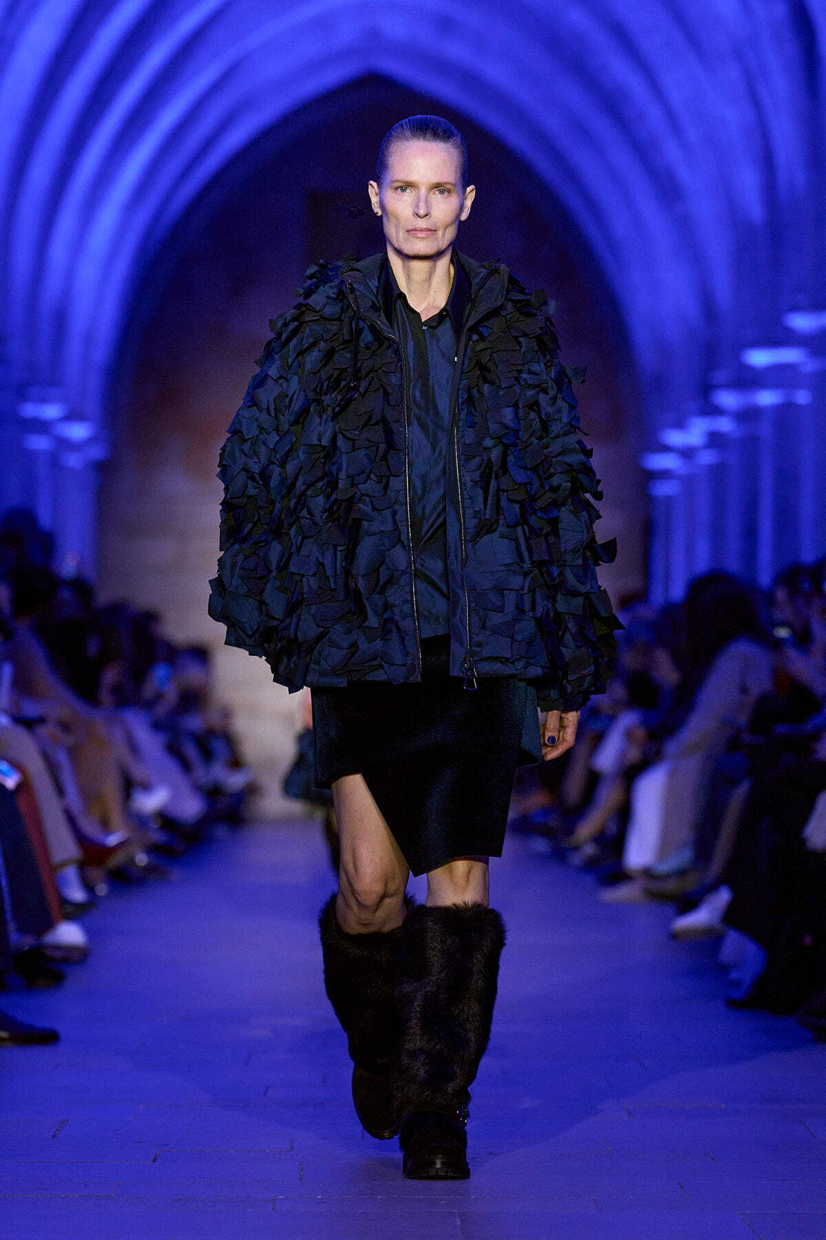 Model walks a blue-lit runway in a voluminous navy textured coat with layered fringe, black skirt, and fur boots.