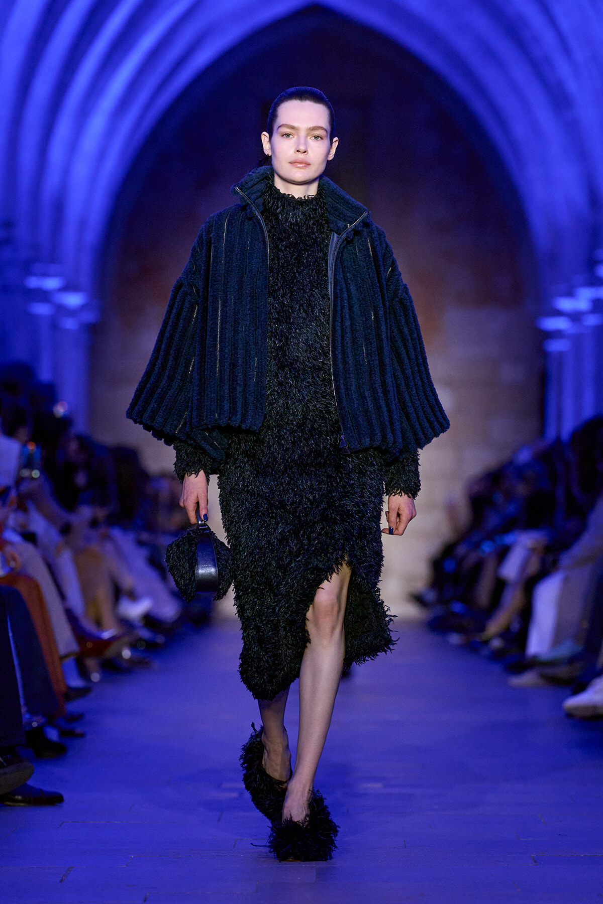 Model walking a runway in a black shaggy dress and oversized blue quilted jacket under blue lighting.