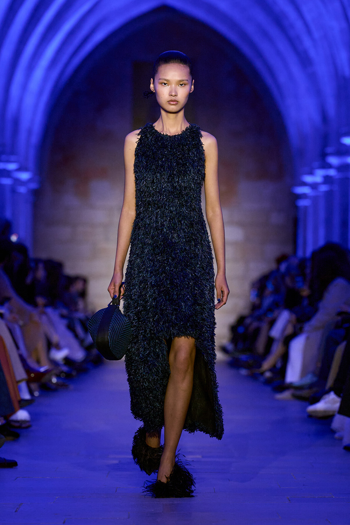 Model walks runway in a sleeveless black textured dress, blue-lit archway background, holding a round striped hat.