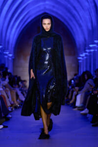 Model walking a blue-lit runway in a sequined dark blue dress and furry coat, under arched blue lighting.
