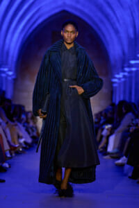 Model on a runway wearing a long navy textured coat over a dark dress, clutch in hand, blue-lit arches in the background.