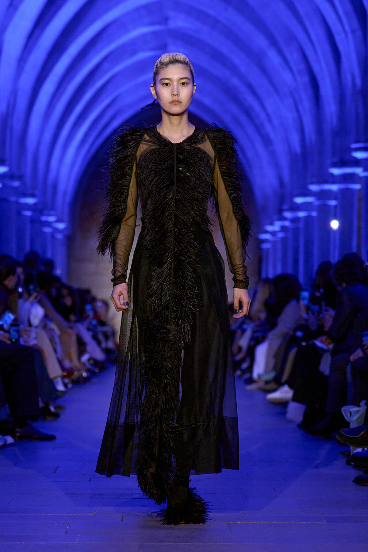 Female model walks a runway in a black feathered gown under blue-lit arches.