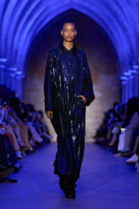 Model walking a fashion runway in a long blue sequined coat under blue archway lighting, hands in pockets, audience on sides