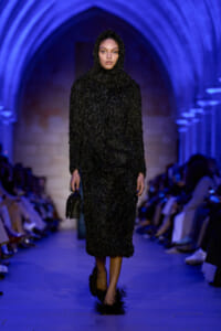 Model walks a blue-lit runway in a full-length black shaggy hooded coat, carrying a small black handbag and feathered shoes visible at the hem.