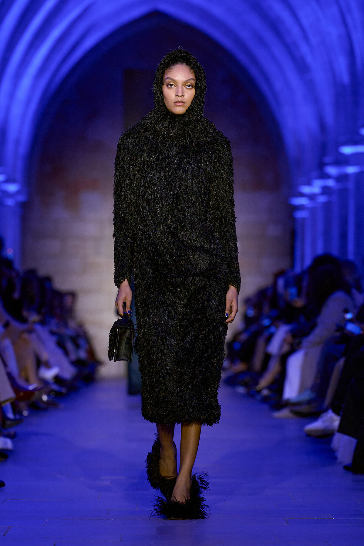 Model walks a blue-lit runway in a full-length black shaggy hooded coat, carrying a small black handbag and feathered shoes visible at the hem.