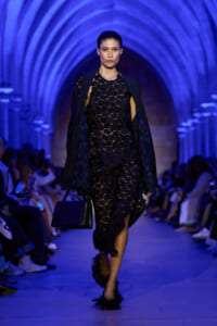 Model walks the runway in a black geometric lattice dress with a cape, carrying a black handbag under blue arched lighting.