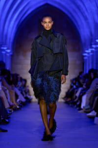 Model walking a runway in a navy trench coat and feathered navy skirt under blue arched lighting.”] ,