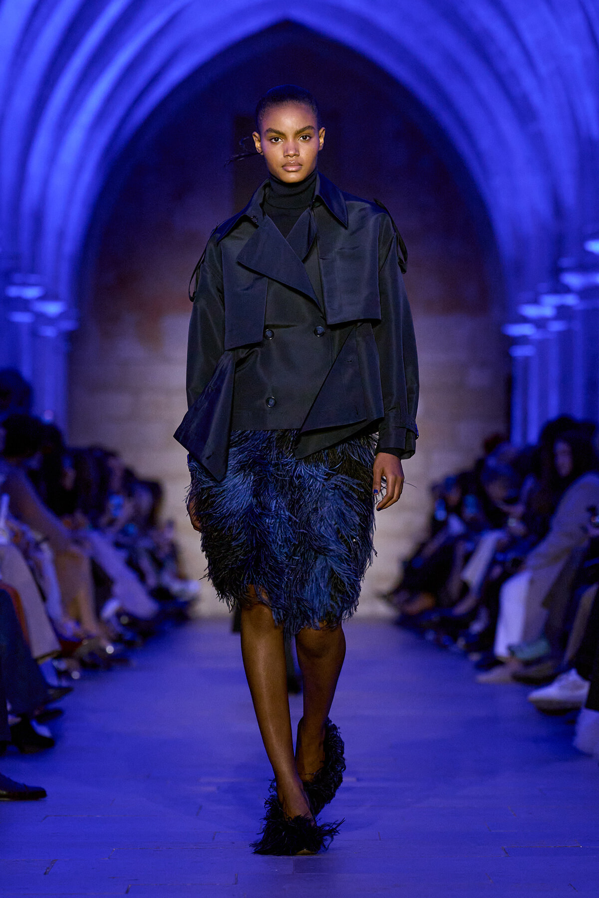 Model walking a runway in a navy trench coat and feathered navy skirt under blue arched lighting.”] ,