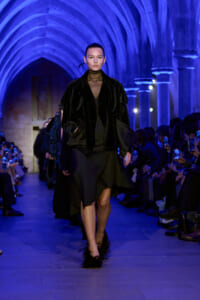 Model walking a dark runway in a black outfit with a voluminous coat and feathered heels, under blue architectural arches during a fashion show.