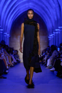 Model walking the runway in a navy dress with black feather trim, blue-lit arched tunnel backdrop.