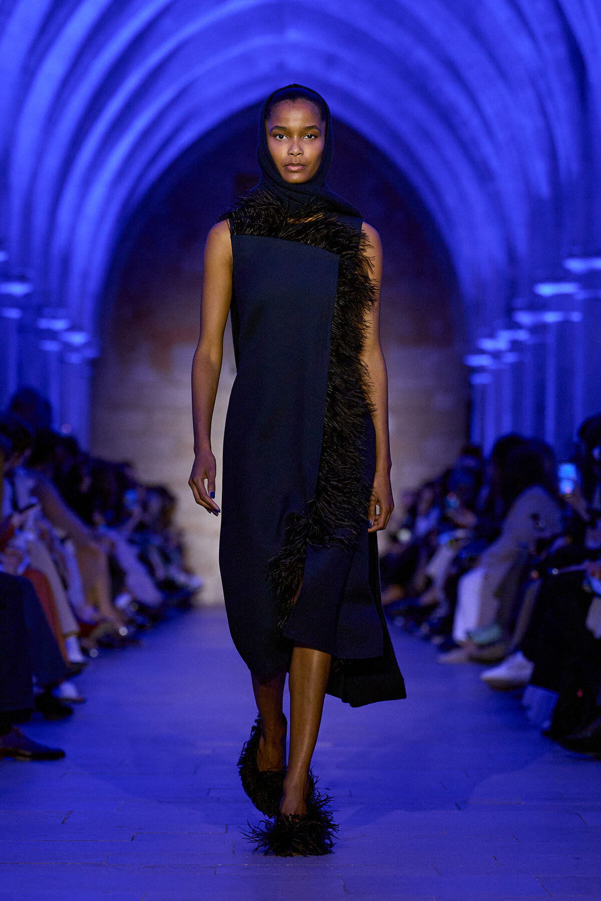 Model walking the runway in a navy dress with black feather trim, blue-lit arched tunnel backdrop.