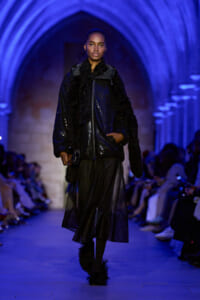 Model walking a fashion runway in a black sequined jacket, sheer black skirt, and feathered boots under blue arched lighting.